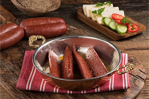 Bakır tavada pişmiş sucuk dilimleri, yanında pastırma, taze sebzeler ve peynirle birlikte sunuluyor.